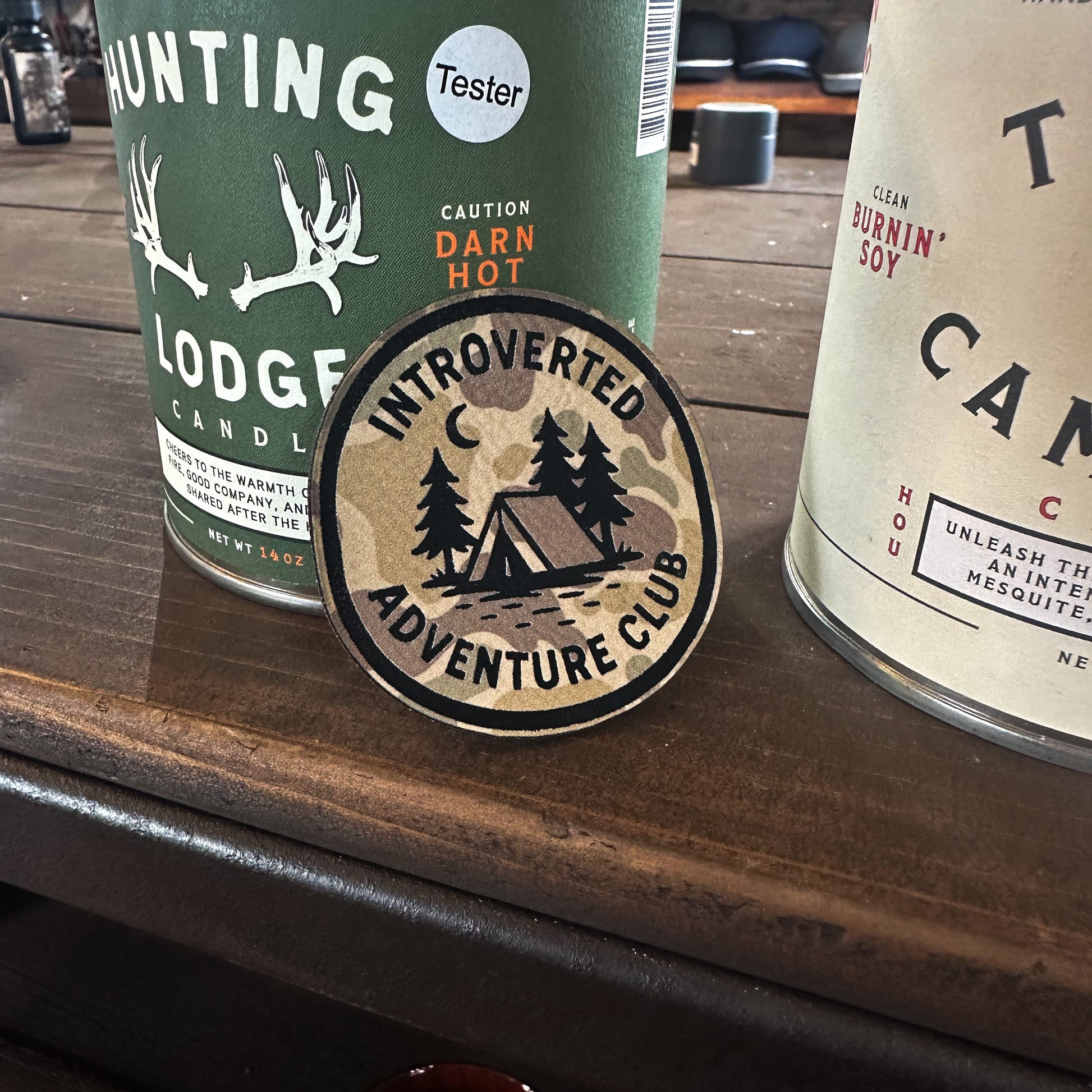 Round sticker with 'Introverted Adventure Club' design on a wooden surface with candles in the background