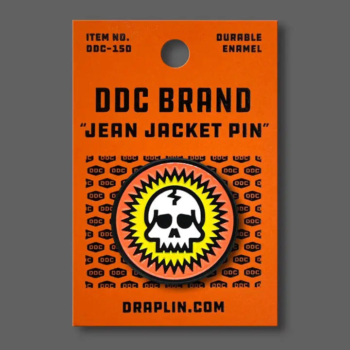 Draplin Design Company skull enamel pin with orange and yellow burst design on orange backing card