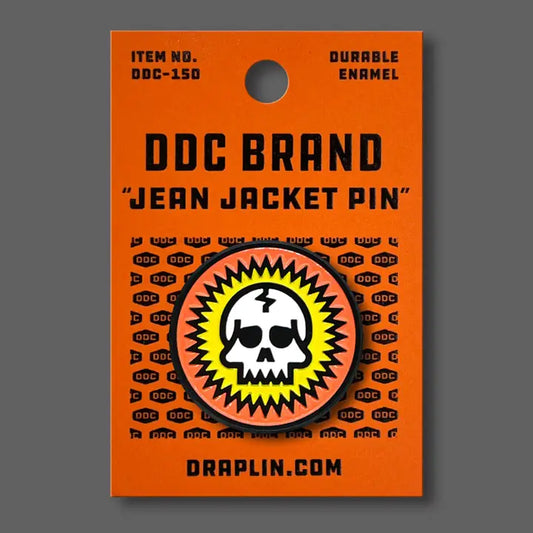 Draplin Design Company skull enamel pin with orange and yellow burst design on orange backing card