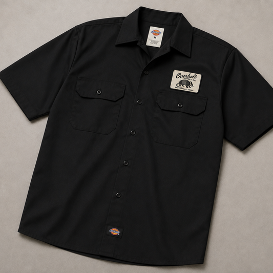 A black short-sleeve button-up work shirt laid flat on a neutral gray background. The shirt features two chest pockets, a 'Overbelt' brand patch on the left chest, and a small orange and blue Dickies logo near the bottom hem.